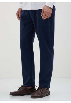 Mann in marineblauer Hose, weißem Hemd und dunkelbraunen Leder-Schnürloafers steht vor einem einfachen hellen Hintergrund.