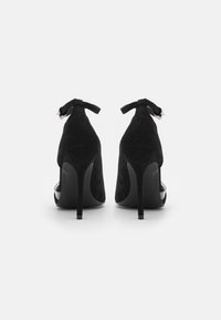 Zapatos de tacón alto de ante negro con punta afilada, correa en el tobillo y un diseño elegante. Los tacones son finos y altos, añadiendo altura.