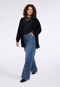 Cardigan nero aperto davanti sovrapposto a una maglia nera a pois. Jeans a vita alta, a gamba larga, di colore azzurro chiaro, e stivali neri con tacco.