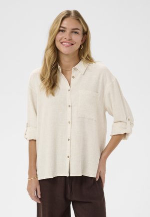 Femeie zâmbitoare cu păr blond lung, purtând o cămașă crem cu nasturi și mâneci suflecate, și pantaloni maro închis, pe un fundal simplu.