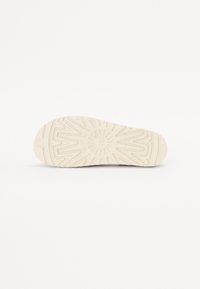 Såle av en sneakers, med en strukturert lysebeige gummioverflate og et mønstret dekke for grep, samt et sirkulært emblem i midten.