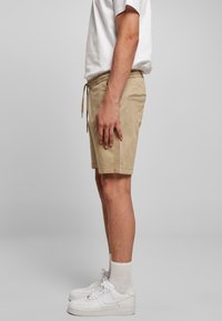 Homme portant un short beige à cordon, des chaussettes blanches montantes, des baskets blanches et un t-shirt blanc, debout de profil devant un fond uni.
