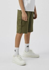 Pantaloni cargo verde oliva con tasche laterali, abbinati a sneaker bianche e calzini bianchi a coste con un piccolo logo nero.