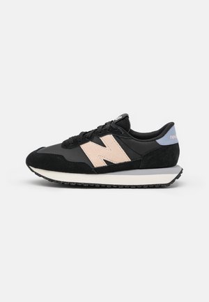 Baskets New Balance noires et beiges avec lacets noirs, semelle texturée et languette de talon bleu clair sur fond blanc.