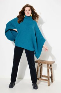 Poncho lavorato a maglia in turchese con colletto alto, caratterizzato da una vestibilità ampia e bottoni decorativi su un lato, abbinato a pantaloni neri e scarpe.