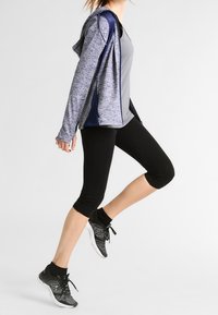 Conjunto deportivo que incluye una camiseta sin mangas gris, una sudadera con cremallera en tonos azul marino y gris, leggings capri negros y zapatillas de running negras texturizadas.