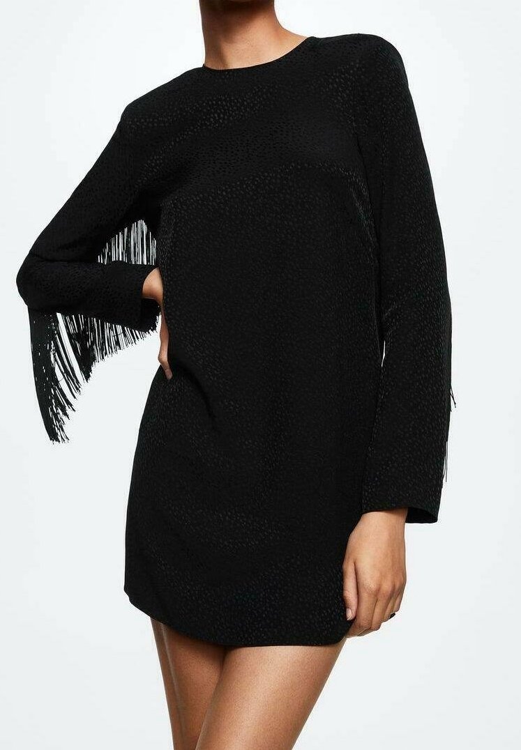 Femme portant une robe courte noire à manches longues avec des détails à franges sur les manches, debout avec une main sur la hanche.