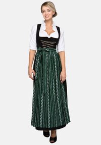 Traditionelles Dirndl, bestehend aus einem schwarzen Mieder mit silberner Schnürung und einem grünen Rock, der mit weißen Blumenmustern verziert ist. Begleitet von einer weißen Bluse.