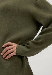 Olivgrüner Strickpullover mit langen Ärmeln, lockerer Passform, gerippten Bündchen und weicher Textur; Hand, die zur Unterstützung an der Seite ruht.