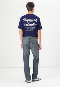 Marineblauwe T-shirt met korte mouwen en witte tekst op de achterkant, gecombineerd met lichtblauwe denim jeans en zwart-witte sandalen.