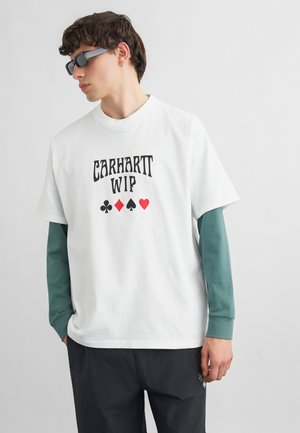 Mladić koji nosi crne sunčane naočale, bijelu Carhartt WIP majicu na slojeve s motivima karata preko dugih zelenih rukava i crne hlače.