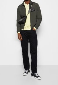 Herrenoutfit mit einer dunkelgrünen Bomberjacke, gelbem Sweatshirt, schwarzen Hosen und schwarzen Sneakers mit weißen Schnürsenkeln. Kleine schwarze Umhängetasche.