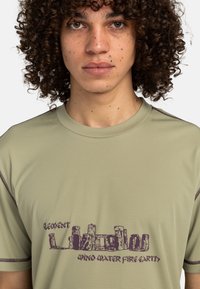 Light green short-sleeve t-shirt with a textured pattern, featuring a purple graphic design depicting stone structures and text.