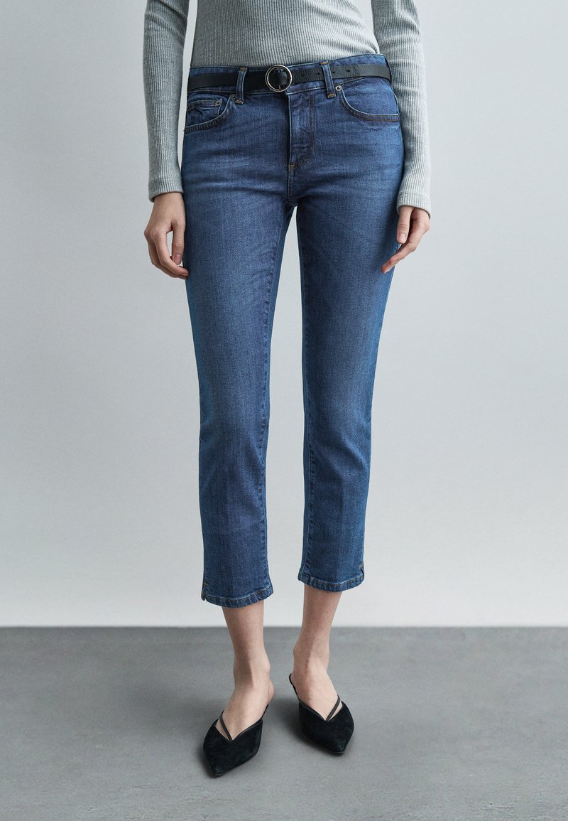 Femme portant un jean bleu raccourci avec une ceinture noire, un haut gris à manches longues côtelé, et des chaussures noires à bout pointu, debout sur un sol gris.