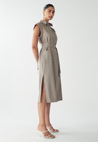 Ärmelloses beiges Kleid mit vertikalen Streifen, tailliertem Bund, seitlichen Schlitzen und zwei vorderen Taschen. Kombiniert mit weißen Absatzsandalen.