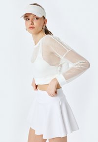 Top corto bianco con maniche lunghe in rete, abbinato a una mini gonnellina bianca a ruota. La modella indossa un berretto abbinato e orecchini a cerchio argentati.