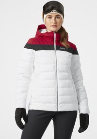 Chaqueta de plumas blanca con bloques de color rojo y negro en los hombros, que cuenta con una cremallera frontal, capucha y puños acanalados. Modelo usando guantes de esquí.