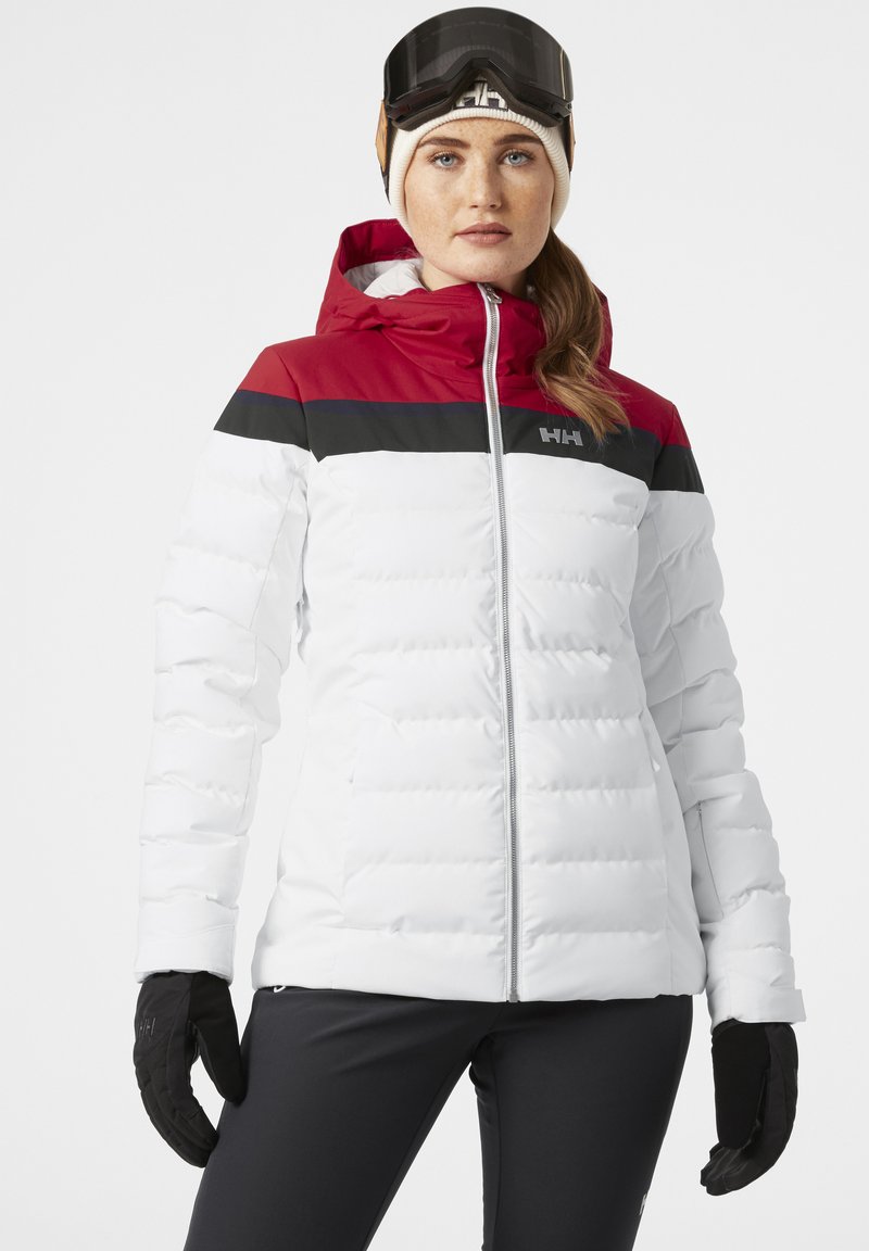 Chaqueta de plumas blanca con bloques de color rojo y negro en los hombros, que cuenta con una cremallera frontal, capucha y puños acanalados. Modelo usando guantes de esquí.