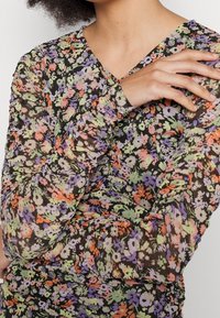 Top de manga larga y escote en V, hecho de tela transparente con un patrón floral multicolor, que presenta detalles fruncidos y un fondo negro.