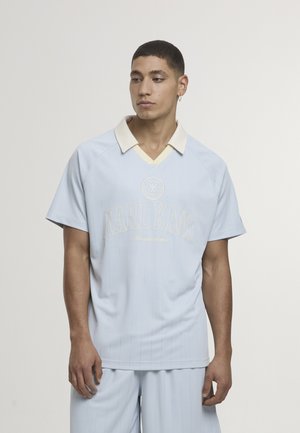 Svetlo modra srajca s kratkimi rokavi in kremno ovratnico, z navpičnimi črtami in napisom "KARL KANI" v beli barvi. Gladek tekstura, sproščen kroj.