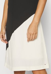 Femme portant une robe à blocs de couleur diagonaux noirs et blancs, avec les mains et les bras visibles, debout devant un fond uni.