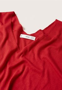 Czerwona dzianinowa bluzka typu v-neck o miękkiej teksturze; zawiera metkę z informacjami o marce. Tkanina wydaje się lekka i lekko układająca się.