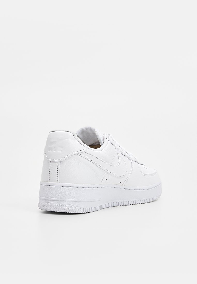Zapatilla baja de cuero blanca con cierre de cordones en la parte delantera, logotipo en relieve en el talón y suela texturizada, vista desde un ángulo trasero sobre fondo blanco.