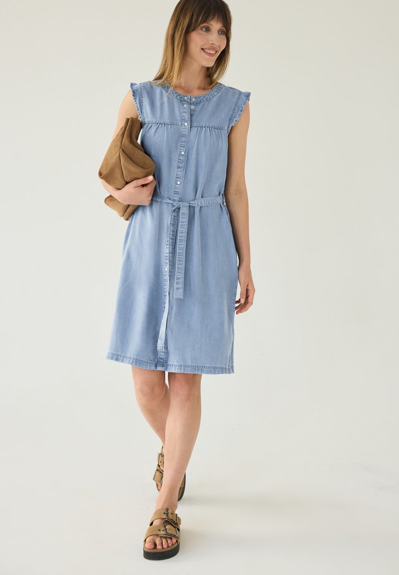 Femme souriante portant une robe sans manches bleu clair boutonnée sur le devant avec ceinture à la taille, tenant une veste marron pliée et des sandales camel à boucle.