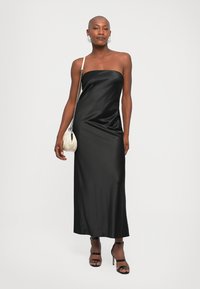 Mujer con un vestido de satén negro sin tirantes, tacones negros, un bolso blanco con cadena y grandes pendientes hoops blancos sobre un fondo liso.