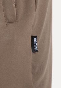 Braune Baumwollhose mit einer Seitentasche und einem schwarzen Etikett mit weißem Text, auf dem der Markenname steht. Glatte Textur und figurbetonter Schnitt.