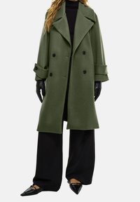 Dobbeltbrystet olivengrøn frakke med brede klapper, sorte knapper og optrillede ærmer, parret med sorte bukser med bred ben og spidse flade sko.