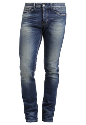 Homme portant une chemise grise à manches longues, un jean bleu, des chaussures marron habillées, portant un sac à dos en cuir marron sur une épaule, marchant vers l'avant.