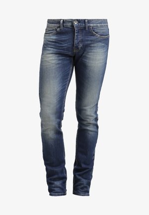 Mann trägt graues Langarmshirt, blaue Jeans, braune elegante Schuhe, trägt braunen Leder-Rucksack über einer Schulter, geht vorwärts.