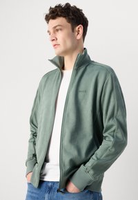 Lys grønn zip-up jakke med et teksturert geometrisk mønster, høy krage og ribbede mansjetter, båret over en hvit T-skjorte og blå jeans.