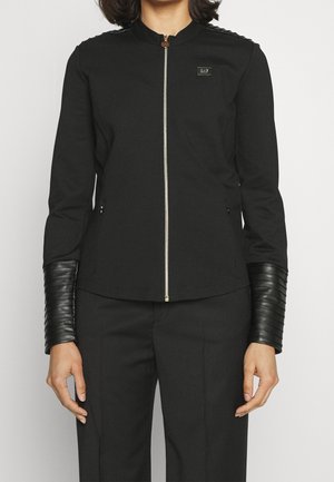 Žena nosící přiléhavou černou bundu se zipem vpředu, s polstrovanými rukávy připomínajícími kůži od loktů až po zápěstí a logem EA7 na hrudi.