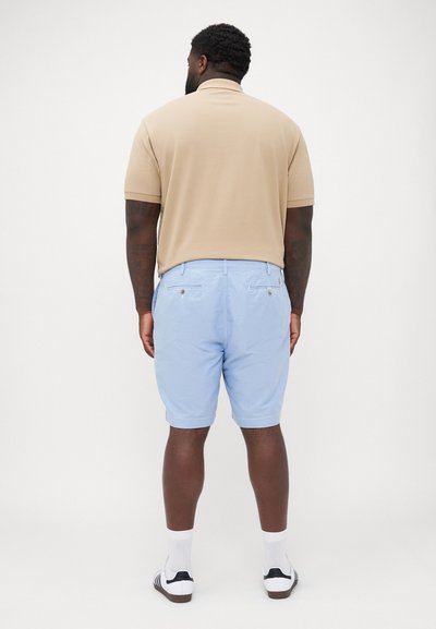 Polo beige à manches courtes, associé à un short bleu clair avec des poches arrière et des détails en doré. Chaussettes blanches mi-mollet et baskets.