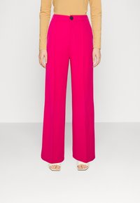 Femme portant un pantalon large taille haute de couleur rose vif, un haut à manches longues beige et des talons carrés blancs ouverts, se tenant devant un fond blanc uni.
