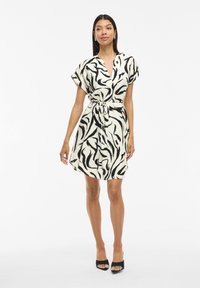 Weißes Kleid mit einem schwarzen abstrakten Muster, kurzen Ärmeln und einem Taillengürtel. Der Stoff wirkt leicht und strukturiert. Getragen mit schwarzen High Heels.
