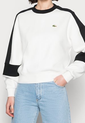 Person iført en hvid sweatshirt med sorte striber på ærmerne og et lille grønt krokodillelogo, kombineret med lyseblå jeans.
