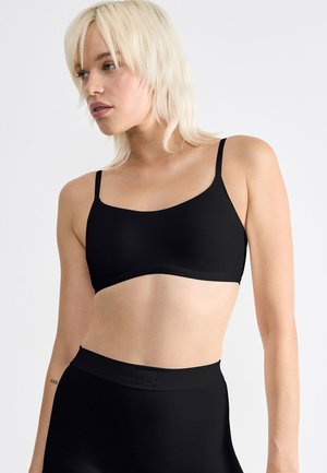 Zwarte draadloze bralette met dunne bandjes, gemaakt van een gladde, rekbare stof en een eenvoudig, naadloos ontwerp. Gecombineerd met hooggeknipte zwarte onderkleding.