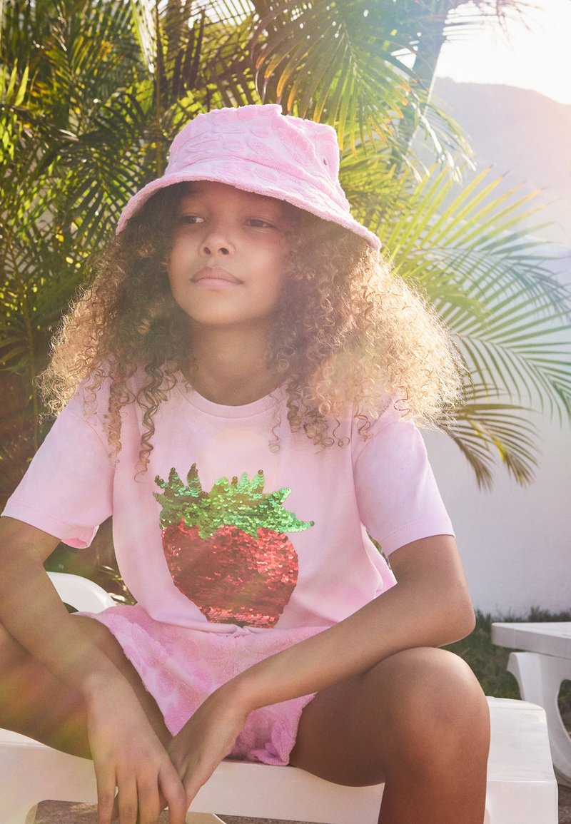 T-shirt di cotone rosa chiaro con un design di fragola in paillettes, abbinata a un cappello a secchiello di un rosa testurizzato. Sullo sfondo ci sono piante tropicali.