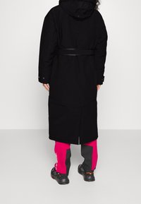 Svart lång kappa med cinchad midja, tillsammans med ljusrosa och gråbyxor. Kappan har huva och knapppdetaljer på ärmarna.