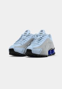Šviesiai mėlyni Nike Shox sportbačiai su tekstūruota viršutine dalimi, perforuotomis detalėmis ir kontrastiniu juodu guminiu padu su mėlynais akcentais.
