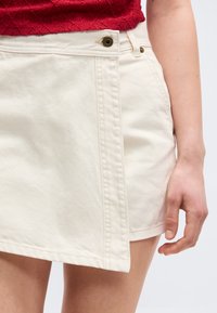 Κοντινό πλάνο ενός ατόμου που φοράει ασύμμετρη κρέμ τζιν φούστα με κουμπί και τσέπη, σε συνδυασμό με ένα υφασμάτινο κόκκινο τοπ.