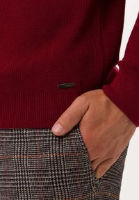 Czerwony sweter wykonany na drutach z ryżowym wykończeniem i mankietami, z małą czarną metalową metką. Połączony z spodniami w kratę w odcieniach szarości, granatu i czerwieni.