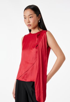 Kobieta o długich czarnych włosach, ubrana w czerwoną satynową bluzkę bez rękawów z drapowanym detalem oraz czarne spodnie, na białym tle.