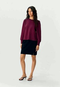 Maroon sweatshirt with puff sleeves, paired with a navy skirt featuring a side slit. Model wearing black flip-flops. Smooth texture.