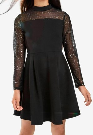 Sort kjole med gennemsigtige lange ærmer og øverste del af brystet prydet med spredte små hvide glimmer, knælang flæse nederdel, båret af en person.