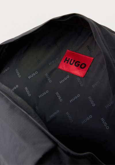 Sac HUGO noir ouvert, laissant voir une doublure intérieure à motifs et une étiquette rouge de la marque HUGO.