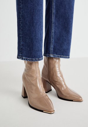 Bottines beige brillant en cuir verni avec talons carrés et bouts pointus en métal, portées avec un jean en denim bleu foncé.